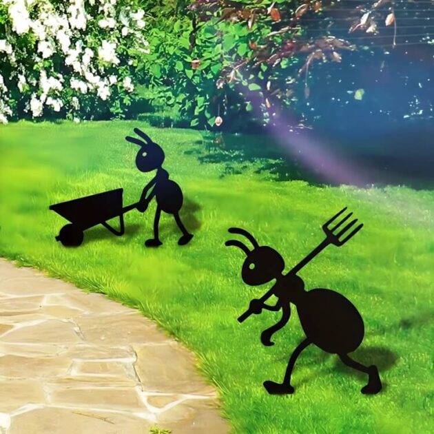Two ant garden figurines with pitchfork and wheelbarrow on a sunny garden lawn near a walking path.