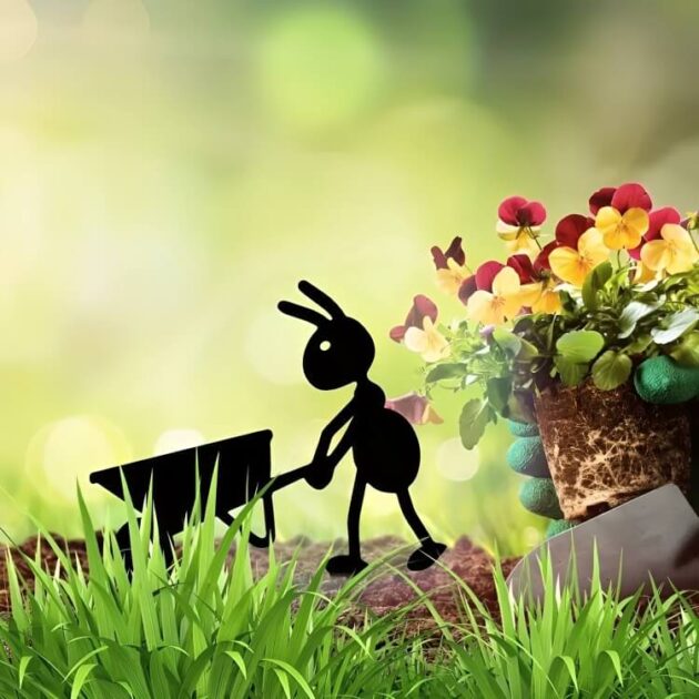 Metal ant statue with a wheelbarrow stands on a stone beside a flowerpot in a green garden.