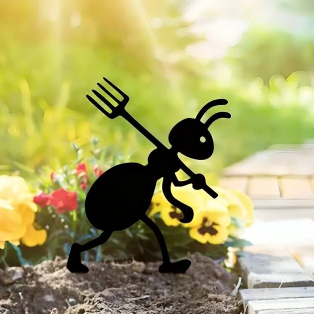 A metal ant with a pitchfork stands among blooming flowers in a garden with a nearby walkway.