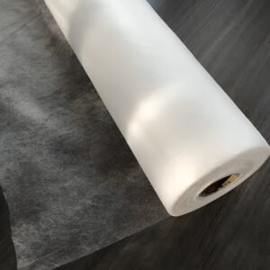 A roll of iron-on fabric hem tape sits on a wooden floor, ready for easy pant hemming at home.